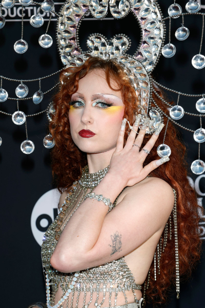 Chappell Roan appears at an event in a glittering headpiece and top. Her nails are covered in newspaper print.